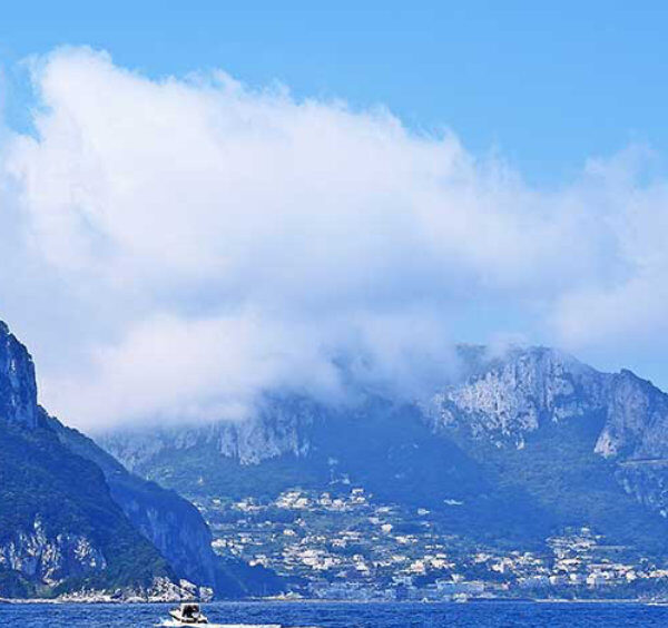 Vue panoramique depuis la mer de l'île de Capri avec ses montagnes rocheuses escarpées, le village côtier niché dans la baie, et de gros nuages blancs dans un ciel bleu.