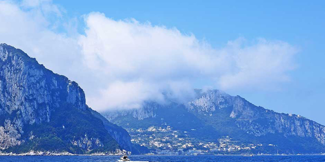 Vue panoramique depuis la mer de l'île de Capri avec ses montagnes rocheuses escarpées, le village côtier niché dans la baie, et de gros nuages blancs dans un ciel bleu.