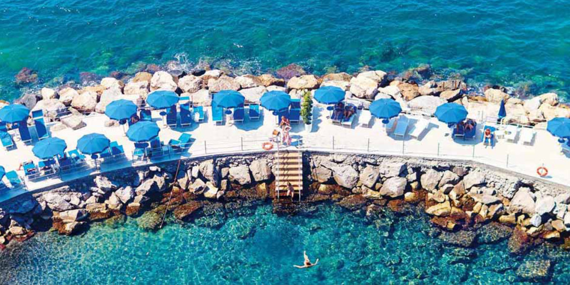 Plage aménagée à Sorrente avec parasols bleus, rochers et eau turquoise.