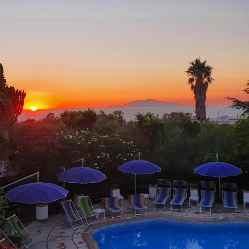 Piscine de la Villa Eva à Capri au coucher du soleil avec parasols bleus et vue sur l'horizon.
