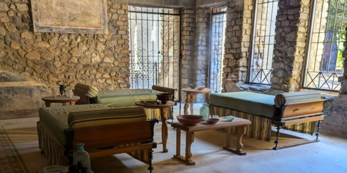 Salle de la Villa Impériale à Pompéi avec lits de banquet romains, petites tables et murs en pierre ancienne.
