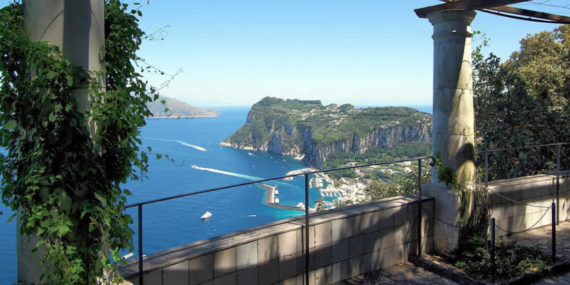 Vue panoramique sur le port et les falaises de Capri depuis la terrasse de la Villa San Michele, encadrée par des colonnes en pierre.