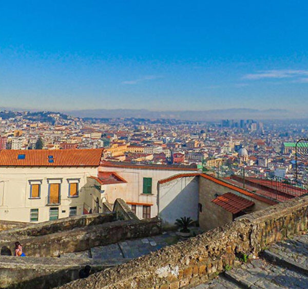 Vue panoramique de Naples depuis une colline avec centre historique, toits et Vésuve en arrière-plan.