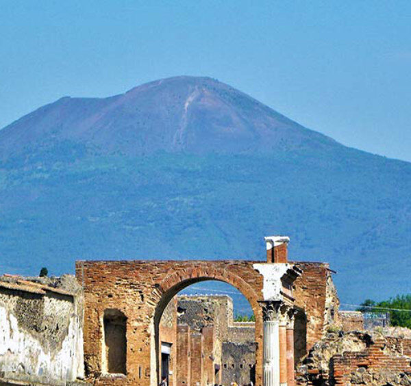 Vue des ruines de Pompéi, incluant un arc en brique et une colonne, avec le mont Vésuve dominant l'horizon.
