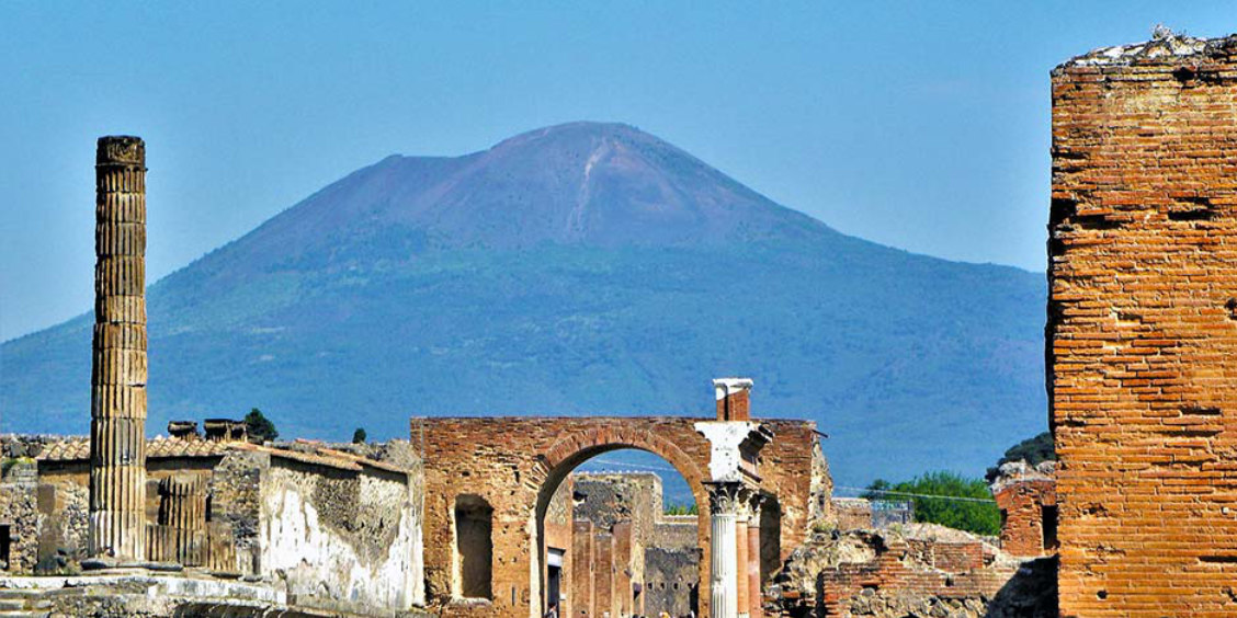 Vue des ruines de Pompéi, incluant un arc en brique et une colonne, avec le mont Vésuve dominant l'horizon.