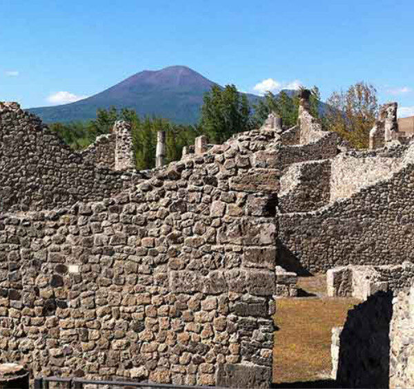 Ruines de Pompéi composées de murets en pierre avec la silhouette du mont Vésuve dominant l'horizon en arrière-plan.
