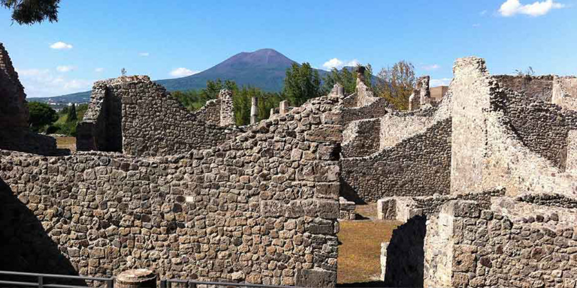 Ruines de Pompéi composées de murets en pierre avec la silhouette du mont Vésuve dominant l'horizon en arrière-plan.