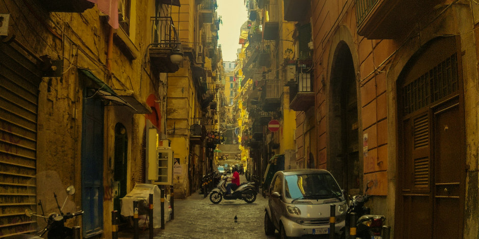 Rue étroite du centre historique de Naples avec voitures, scooters et ambiance urbaine typique.