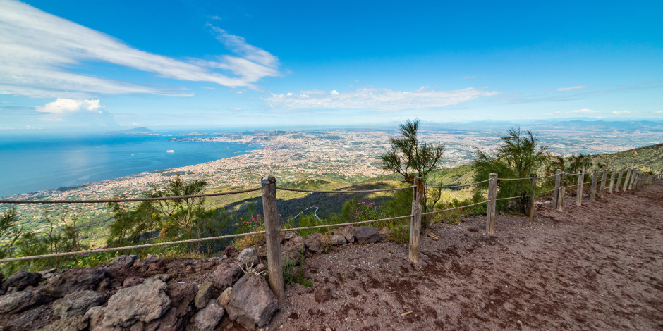 Vue panoramique de la baie de Naples et du littoral depuis le sommet du Vésuve, avec un sentier de randonnée bordé d'une clôture en bois au premier plan.