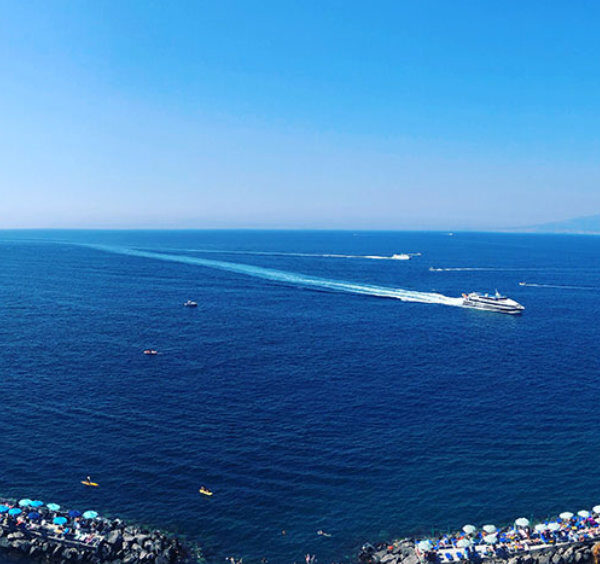 Vue panoramique sur la mer à Sorrente avec bateaux, plages aménagées et Vésuve à l’horizon.