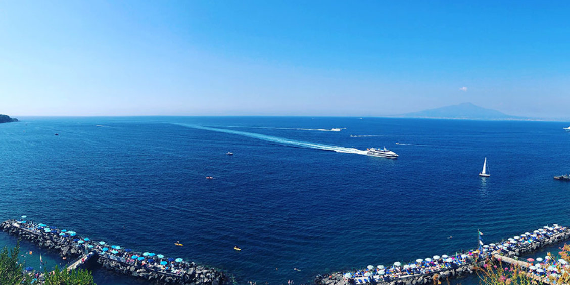 Vue panoramique sur la mer à Sorrente avec bateaux, plages aménagées et Vésuve à l’horizon.